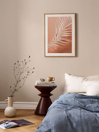 Gerahmtes Poster mit Palmblatt in warmen Beige- und Terrakottatönen über einem Bett.