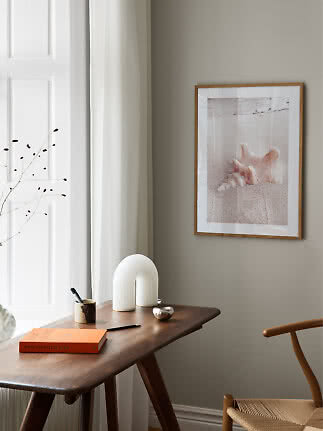 Framed beach print with seashell on sand above a dark wooden desk in a bright room