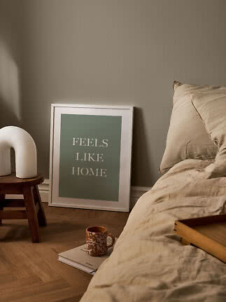 Cozy bedroom with beige bedding, framed poster on the floor, white lamp on stool, and mug on book
