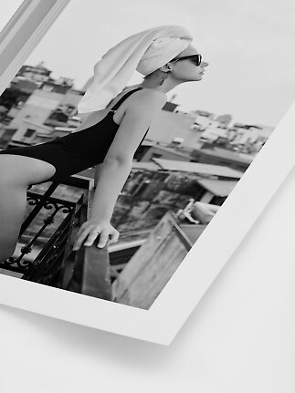 Black-and-white photo of woman in black swimsuit and sunglasses with towel-wrapped hair on balcony
