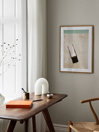 Framed art print of a chair on a beige background above a wooden desk in a bright minimalist room