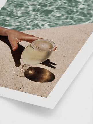 Hand reaching toward a glass of pale drink beside a sunlit pool