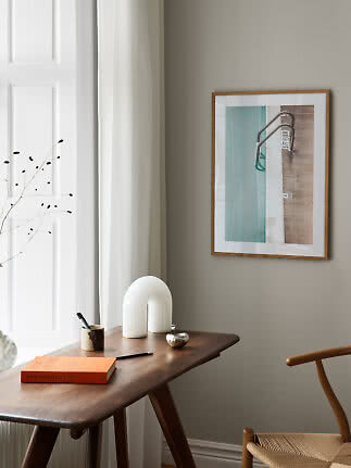 Wooden desk with book, small cup, white sculptural lamp and framed art on a gray wall