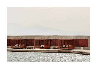Wooden cabanas with sun loungers by a pool, against a shuttered wall and misty mountains