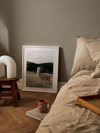 Gemütliches Schlafzimmer mit beiger Bettwäsche, Holzschemel, weißer Lampe und gerahmtem Alpaka-Poster