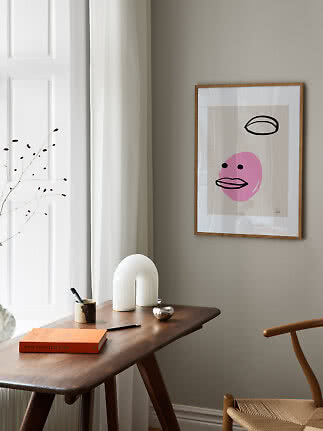 Framed abstract poster with a pink face hanging above a wooden desk in a bright room