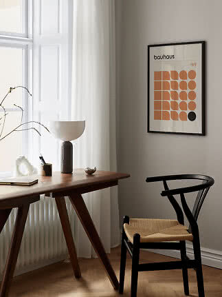 Bauhaus-plakat i sort ramme over træskrivebord med lampe og sort stol ved vindue