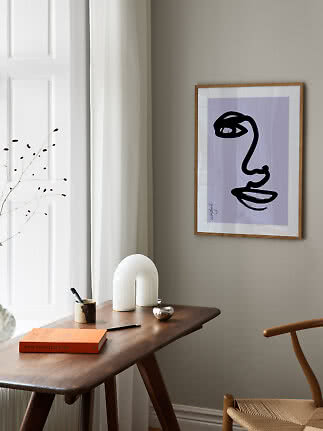 Framed abstract face print on a beige wall above a wooden desk by a window