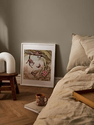 Cozy bedroom with beige bedding, table lamp, and framed jungle print leaning on the floor