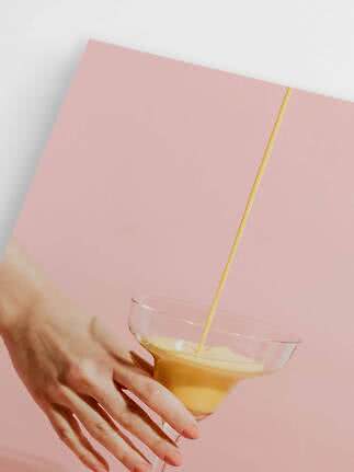 Main tenant un verre à cocktail pendant qu'une boisson orange crémeuse est versée sur fond rose