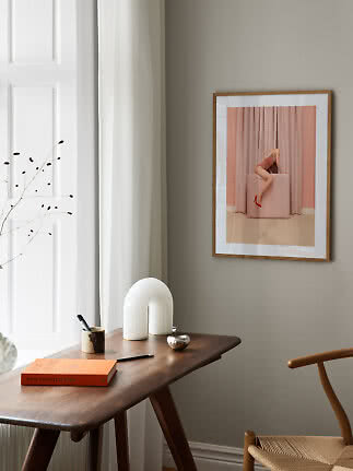 Inramad konstbild med kvinna på rosa kub framför rosa gardiner på beige vägg