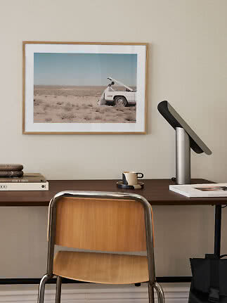 Minimalistisk skrivebord med træstol, lampe, bøger, kop og indrammet ørkenfoto på væggen