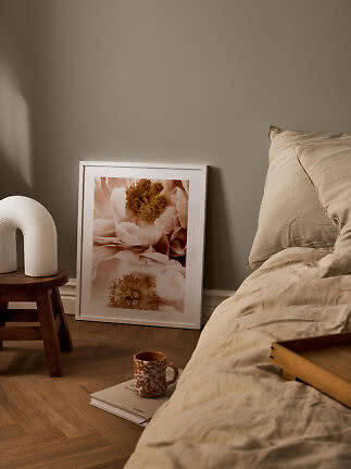 Soveværelse med beige sengetøj, blomsterplakat i hvid ramme og hvid lampe på træskammel