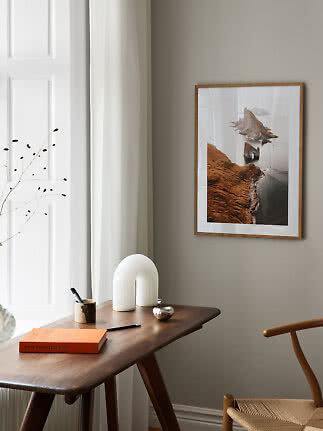 Framed coastal landscape print on wall above wooden desk in a bright minimalist room