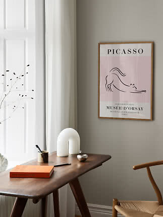 Framed Picasso poster with pink cat illustration above a wooden desk by a window