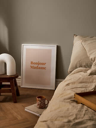 Hyggeligt soveværelse med beige sengetøj, indrammet Bonjour Madame-plakat og hvid lampe på skammel