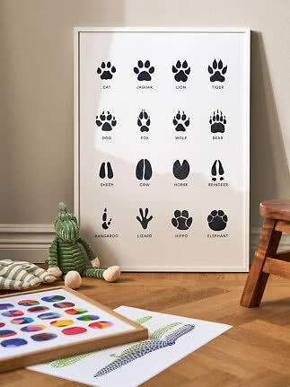 White framed poster with black animal footprints and labels, leaning against a nursery wall