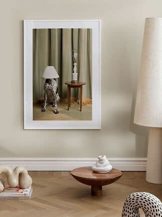 Indrammet kunsttryk af plettet hund med lampeskærm på hovedet ved siden af et lille bord med lampe