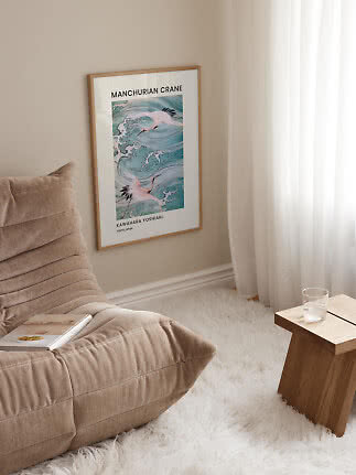 Inramad konstposter med tranor över blå vågor på beige vägg bredvid fåtölj och gardin