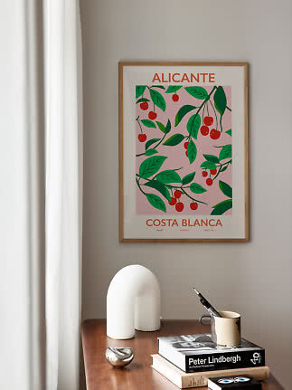 Indrammet Alicante-plakat med kirsebærmotiv hænger over et træbord med bøger og kop