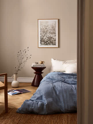 Gemütliches Schlafzimmer mit blauer Bettdecke, weißen Kissen, dunklem Beistelltisch und Blumenbild