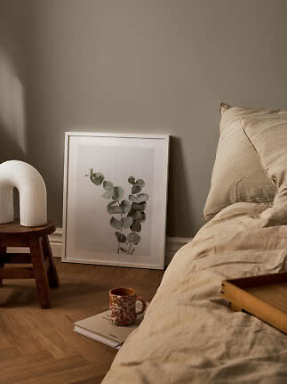 Hyggeligt soveværelse med beige sengetøj, botanisk plakat, hvid lampe på skammel og krus på bog