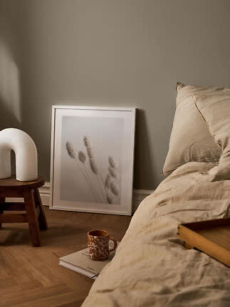 Gemütliches Schlafzimmer mit beiger Bettwäsche, gerahmtem Pflanzenbild und weißer Lampe auf Hocker
