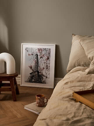 Gemütliches Schlafzimmer mit beiger Bettwäsche, Tischlampe und gerahmtem Eiffelturm-Poster am Boden