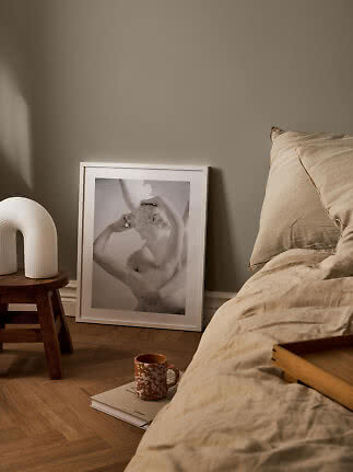 Hyggeligt soveværelse med beige sengetøj, indrammet kunst, hvid bordlampe og krus på bog