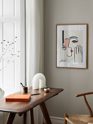 Póster enmarcado con rostro abstracto en pared beige sobre mesa de madera en una estancia luminosa