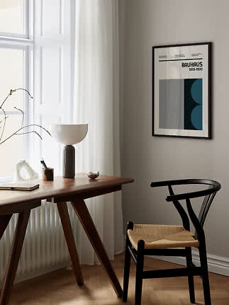 Plakat Bauhaus w czarnej ramie na jasnej ścianie nad biurkiem i czarnym krzesłem