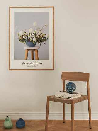 Inramad blomsterposter på beige vägg ovanför trästol med böcker och blå vas