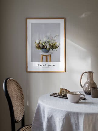 Inramad blomsterposter ovanför bord med kopp, kanna och kaka i mjukt solljus