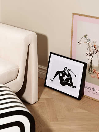 Sort indrammet abstrakt kunsttryk lænet mod væg ved cremefarvet stol og blomsterplakat