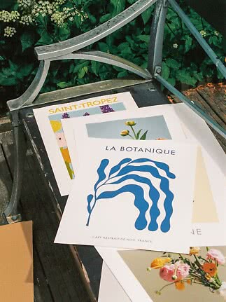 Stampe d’arte e poster floreali su una panca da giardino sotto un bracciolo in metallo.