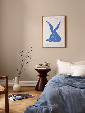 Framed art print with blue figure on beige bedroom wall above bed and dark side table