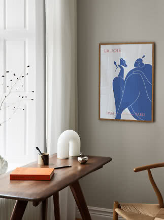 Wood-framed art poster with blue figure on a beige wall above a wooden desk by a window