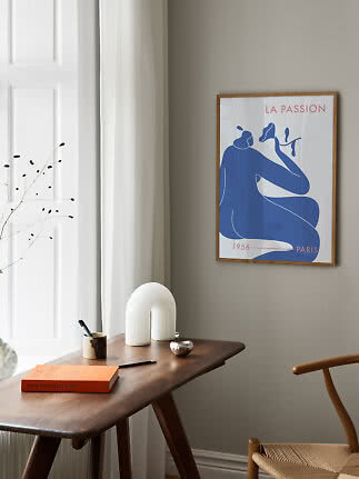 Framed art print with blue abstract figure above a wooden desk in a bright room corner