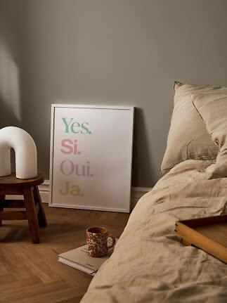 Plakat med ordene "Yes. Si. Oui. Ja." læner op ad væggen ved en seng i et varmt soveværelse