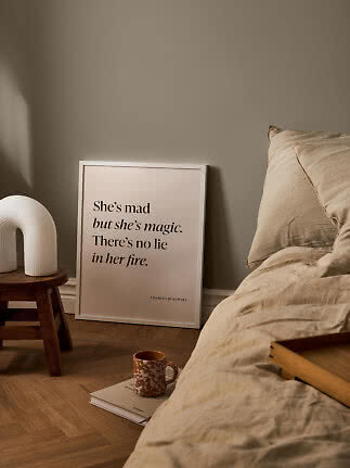 Plakat med citat i hvid ramme lænet op ad væggen ved seng, skammel og kop på bog