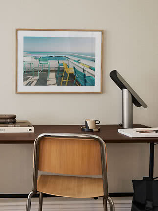 Stampa artistica incorniciata sopra scrivania con sedia, lampada e tazza in studio minimalista