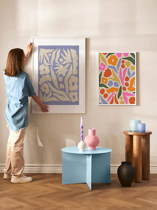 Frau richtet zwei farbige Blumenposter an einer Wand in einem hellen Raum aus.