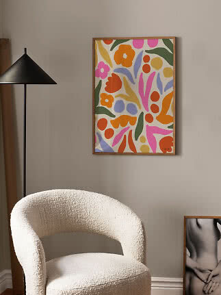 Póster floral abstracto colorido en marco de madera sobre pared beige, encima de un sillón crema.