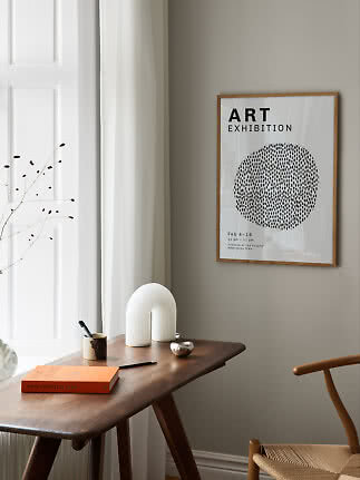Framed art poster above a wooden desk with an orange book, pen, and white decorative lamp