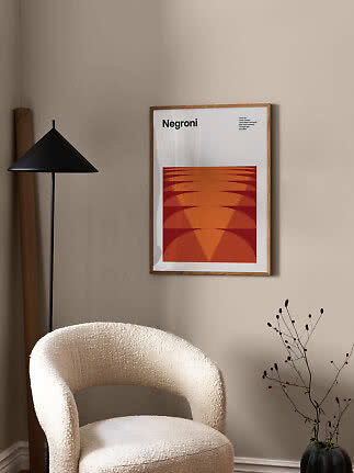 Gerahmtes Negroni-Poster mit orangefarbener Grafik an beigefarbener Wand über weißem Sessel