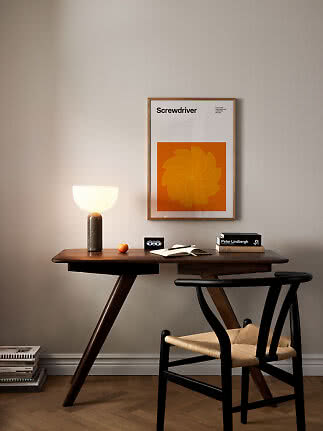 Dunkler Holzschreibtisch mit schwarzem Stuhl, leuchtender Tischlampe und orangefarbenem Poster.