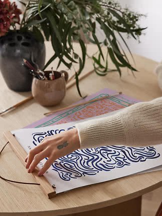 Hand placing art print on wooden table beside vase with greenery and paintbrushes
