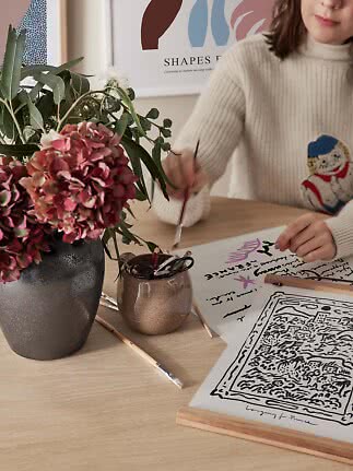 Person painting at a table with flowers in a dark vase, brushes and art prints.