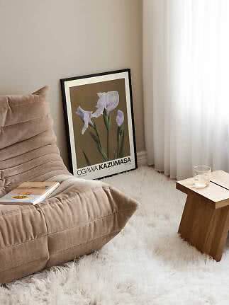Indrammet blomsterplakat med lyse lilla iris lænet mod væg ved beige lænestol og lille træbord