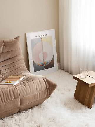 Inramad konstposter lutad mot väggen vid beige fåtölj, vit matta och litet träbord